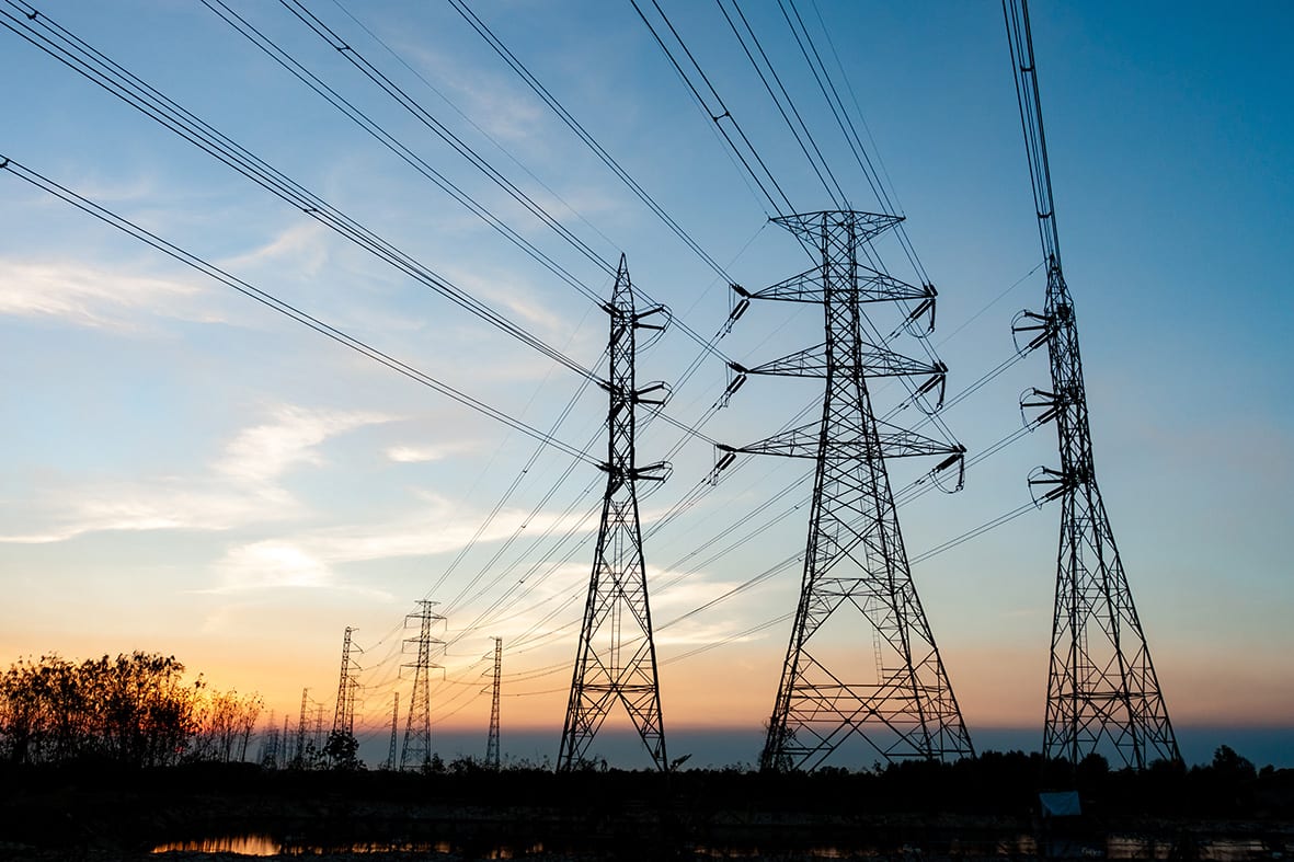A line of high-voltage transmission pylons carrying power lines across open ground at dusk